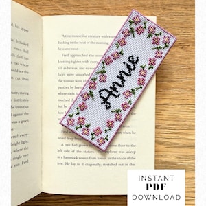 Könnte beinhalten: Handgefertigter Kreuzstich-Lesezeichen mit dem Namen "Annie" in schwarzer Schrift. Das Lesezeichen ist weiß mit einem Rand aus rosa Blumen und grünen Blättern. Unten steht "INSTANT PDF DOWNLOAD".