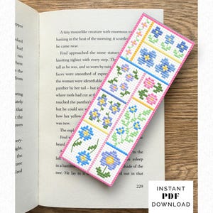 May include: A handmade bookmark with a floral cross-stitch design. The bookmark features a white background with blue, purple, and pink flowers in a grid pattern, framed by a pink border. The bookmark is resting on an open book.