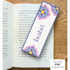 Könnte beinhalten: Ein rechteckiges Kreuzstich-Lesezeichen mit weißem Hintergrund und violettem Rand. Das Design zeigt florale Motive in Pink, Blau und Gelb an den oberen und unteren Ecken. Der Name Isabel ist in der Mitte in Violett gestickt.
