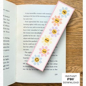 May include: A rectangular cross-stitch bookmark featuring a vertical row of six pastel-colored flowers in blue, peach, lavender, yellow, and pink. Each flower center contains a black letter, spelling out the name LIVIA vertically on the bookmark.