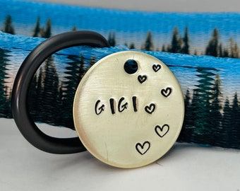 Peut inclure: Une médaille de chien dorée avec le nom "GIGI" gravé dessus. La médaille est attachée à un anneau en métal noir et comporte plusieurs petits cœurs gravés sur le bord.
