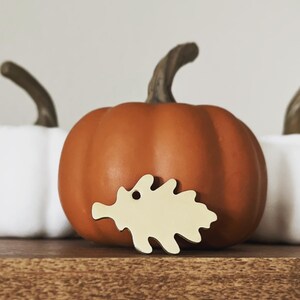 Puede incluir: Un pequeño adorno de madera en forma de hoja de color amarillo claro se encuentra delante de una calabaza naranja. La calabaza está sobre una superficie de madera.