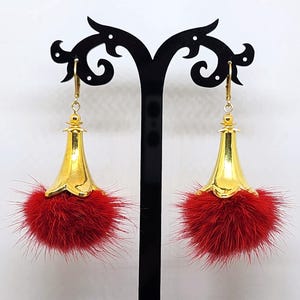 May include: A pair of gold-colored earrings with a bell-shaped design and red fluffy accents. The earrings are suspended from a gold-colored hook. The earrings are displayed on a black stand against a white background.