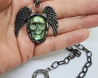May include: A silver-toned necklace featuring a skull pendant with wings. The skull has a green and yellow iridescent appearance. The necklace chain has a toggle clasp. The pendant is approximately 2 inches wide.