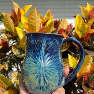 May include: A handmade ceramic mug with a unique blue and green glaze. The mug features a textured design and a curved handle. The mug is approximately 4 inches tall and is perfect for coffee or tea.