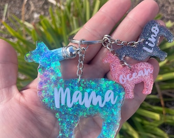 May include: A set of three glitter llama keychains. The largest keychain is blue and says "Mama". The medium keychain is pink and says "Ocean". The smallest keychain is gray and says "Lino".