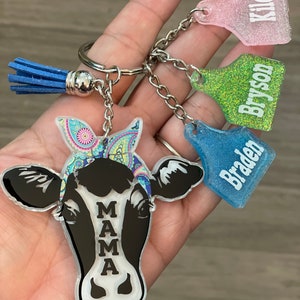 May include: A black and white cow keychain with a colorful bandana. The keychain has a silver ring and a blue tassel. There are three additional keychains with the names "Kiley", "Bryson", and "Braden" on them.
