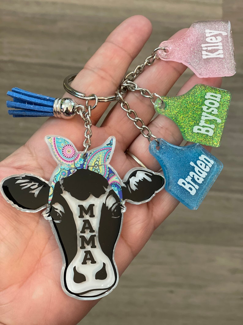 May include: A black and white cow keychain with a colorful bandana. The keychain has a silver ring and a blue tassel. There are three additional keychains with the names "Kiley", "Bryson", and "Braden" on them.