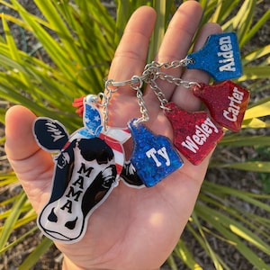 May include: A set of five keychains featuring a cow with a red, white, and blue bandana, and four other keychains with glitter and the names "Ty", "Wesley", "Carter", and "Aiden".