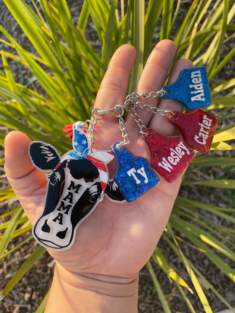 May include: A set of five keychains featuring a cow with a red, white, and blue bandana, and four other keychains with glitter and the names "Ty", "Wesley", "Carter", and "Aiden".
