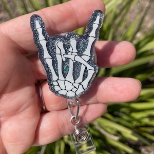 May include: A black and white skeleton hand making the rock and roll hand gesture. The hand is on a black glitter background and is attached to a clear plastic retractable badge holder.