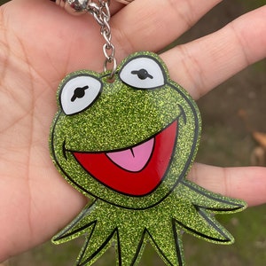 May include: Green glitter keychain featuring a cartoon frog with a wide open mouth and a pink tongue. The frog has black eyes and a black outline.