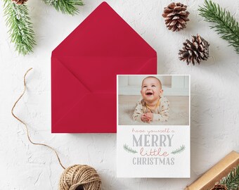 May include: A red envelope and a Christmas card with a baby's photo and the text "Have yourself a Merry Little Christmas." The card is surrounded by pine cones, greenery, and twine on a white surface.