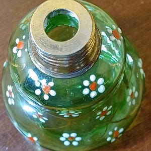 May include: A green glass bottle stopper with a metal rim and screw threads. The stopper is decorated with small white flowers with orange centres. The glass has a slight swirl design.