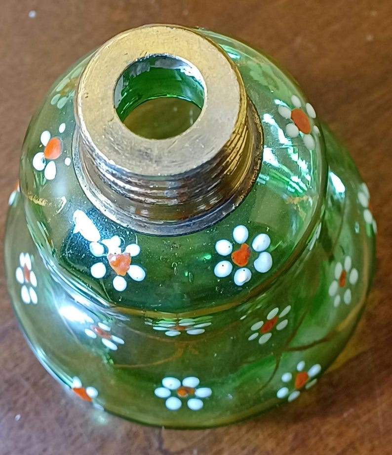 May include: A green glass bottle stopper with a metal rim and screw threads. The stopper is decorated with small white flowers with orange centres. The glass has a slight swirl design.