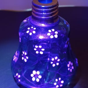 May include: A blue glass bottle with a floral pattern. The bottle has a flared base and a threaded neck. White flowers are painted on the surface, and the bottle has a gold-coloured rim.