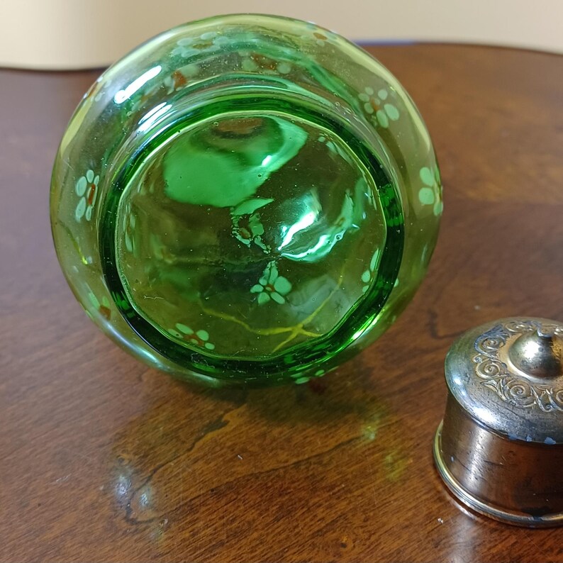 May include: A green glass bottle with a floral pattern and a silver-coloured lid. The bottle is round and has a slightly textured surface. The lid is small and has an ornate design.