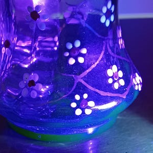 May include: A clear glass vase with a flared base and a rounded body. The vase is decorated with white and red floral designs and green accents. The vase is illuminated with a purple light.