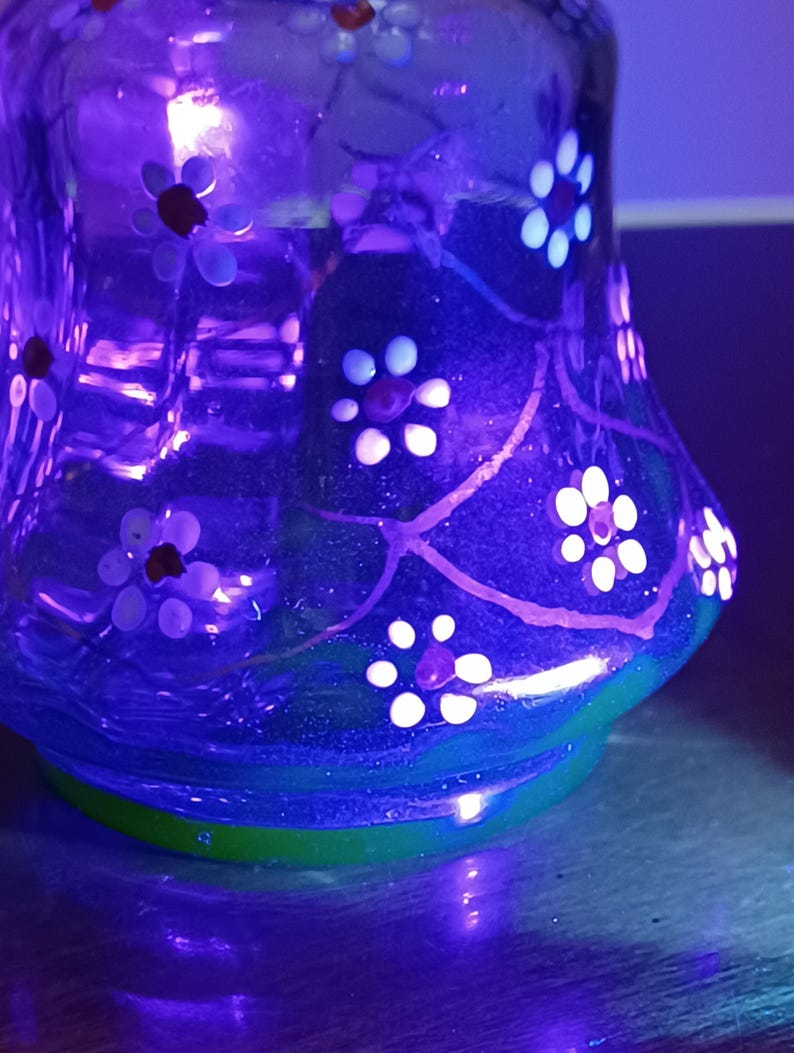 May include: A clear glass vase with a flared base and a rounded body. The vase is decorated with white and red floral designs and green accents. The vase is illuminated with a purple light.