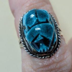 May include: A silver ring featuring a turquoise scarab beetle design. The beetle is intricately carved with detailed wings and body segments. The ring has a braided silver border and a vintage aesthetic.