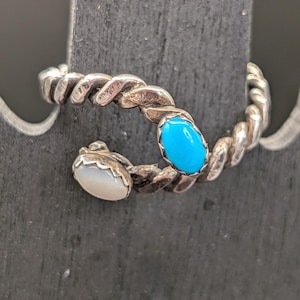 May include: A bypass ring featuring a twisted silver band with an open design. One end is set with an oval, bright blue turquoise stone, while the other end features a smooth, oval white stone in a scalloped silver setting.