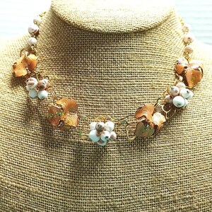 May include: A gold-toned necklace featuring floral and beaded elements. The necklace has copper-colored flower-shaped pendants and clusters of white and brown beads. The necklace is displayed on a burlap-textured surface.