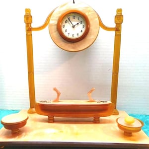 May include: A vintage amber Bakelite clock with a round face and black hands. The clock is mounted on a stand with two small amber bowls on either side. The clock face has black Roman numeral markings and the words 'Made in England' are visible on the clock face.