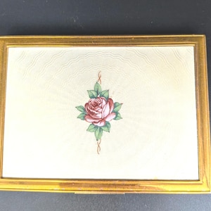 May include: A rectangular gold-toned compact with a cream-colored surface. The surface features a rose design in shades of red, pink, and green. The compact has a gold frame and a textured pattern on the cream surface.