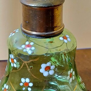 May include: A vintage green glass perfume bottle with a brass cap. The bottle is decorated with white and orange floral designs and gold-coloured vines. The bottle has a rounded base and a flared shape.