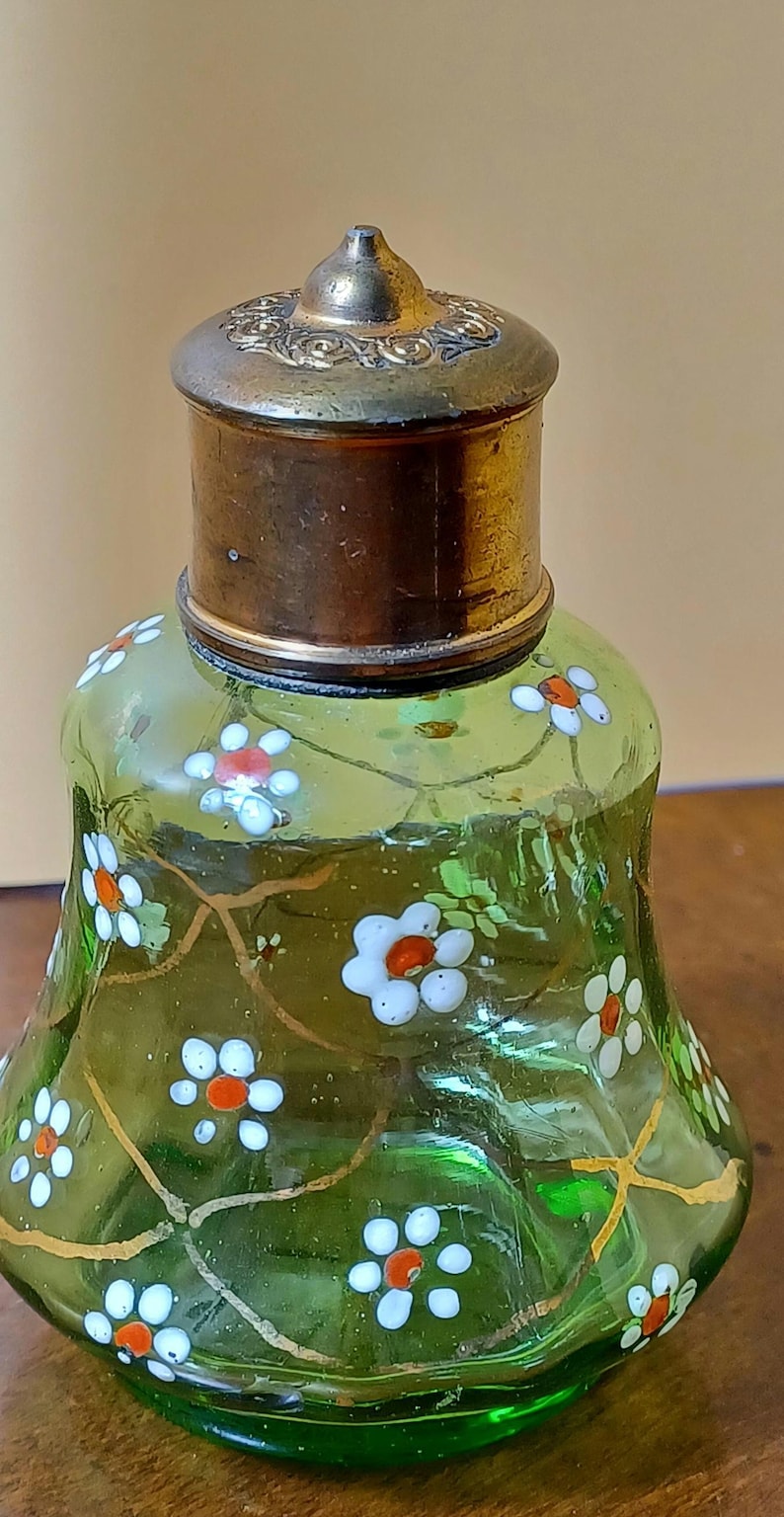 May include: A vintage green glass perfume bottle with a brass cap. The bottle is decorated with white and orange floral designs and gold-coloured vines. The bottle has a rounded base and a flared shape.