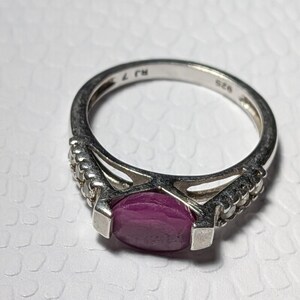 May include: A silver ring with a large oval, deep red gemstone as the focal point. The ring has a decorative band with small, clear stones on each side of the main stone. The band is marked with "925" and other markings.