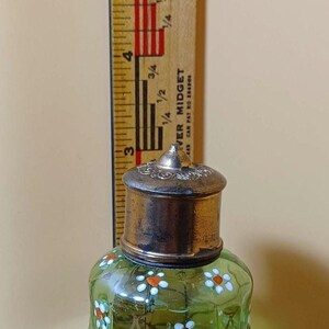 May include: A green glass perfume bottle with a silver-colored metal lid, decorated with white and orange floral designs. A wooden ruler with inch markings is next to the bottle. The bottle is approximately 7.6 cm tall.