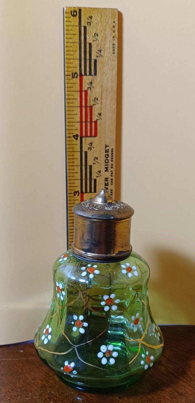 May include: A green glass perfume bottle with a silver-colored metal lid, decorated with white and orange floral designs. A wooden ruler with inch markings is next to the bottle. The bottle is approximately 7.6 cm tall.