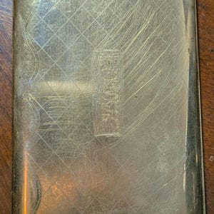 May include: A rectangular, antique metal cigarette case with a textured, scratched surface. The case has a diamond pattern etched into the metal, with a rectangular emblem in the center. The case is a muted gold color.