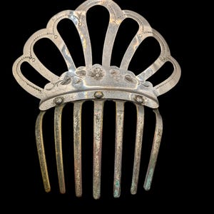 May include: A silver-toned metal hair comb with a crown-like design. The comb features a fan-shaped top with floral and dot details, and vertical tines. The metal has a slightly aged appearance.