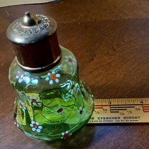 May include: A vintage green glass perfume bottle with a silver-coloured lid and a dark brown neck. The bottle is decorated with hand-painted white and red flowers. A wooden ruler is next to the bottle.