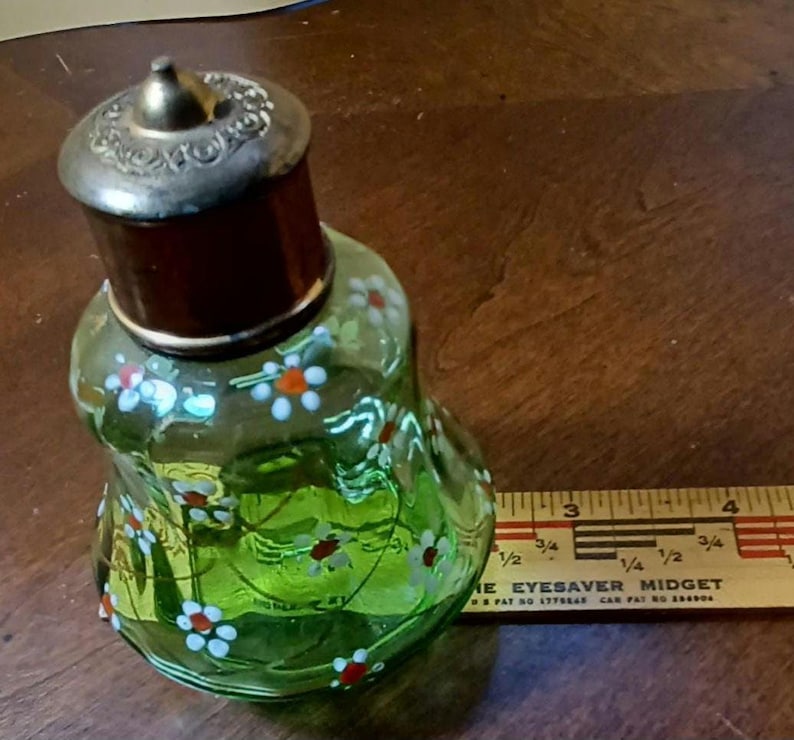May include: A vintage green glass perfume bottle with a silver-coloured lid and a dark brown neck. The bottle is decorated with hand-painted white and red flowers. A wooden ruler is next to the bottle.