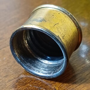 May include: A vintage brass napkin ring with a tarnished, aged appearance. The ring has a cylindrical shape with a slightly flared opening. The interior shows signs of wear, adding to its antique charm. The brass has a warm, golden hue.