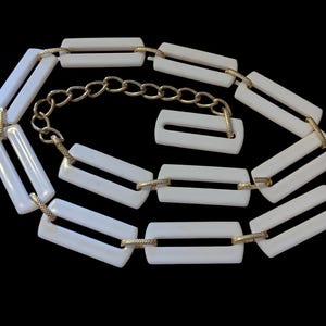 May include: A white rectangular belt with gold-tone metal accents and a chain closure. The belt features a series of white, rectangular links connected by gold-colored metal. The belt is set against a black background.