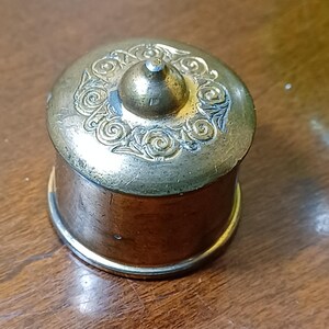 May include: A small, antique brass box with a decorative lid. The lid features an ornate floral design and a small knob. The box has a cylindrical shape with a copper-coloured body and a brass rim.