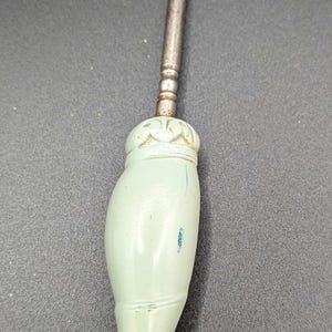 May include: A vintage shoe-shaped button hook with a metal hook and shaft. The handle is a light green color, shaped like a shoe, and has a decorative top. The hook is curved.