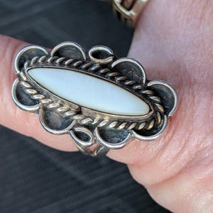 May include: A silver ring with an oval, white mother-of-pearl center stone. The ring has a scalloped edge and a twisted rope design around the stone. The ring is on a finger.