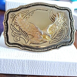 May include: A gold-colored rectangular belt buckle with a detailed eagle design. The eagle is in flight, with outstretched wings, and is the focal point of the buckle. The buckle has a silver border with a decorative pattern.