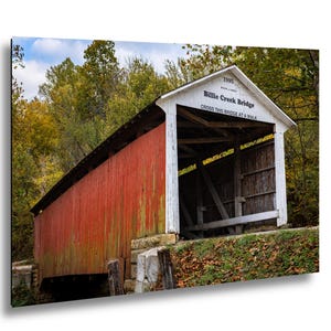 Op de afbeelding: Een ingelijste print van de Billie Creek Bridge, een overdekte brug met rode gevelbekleding en een wit zadeldak. De brug is omgeven door bomen met herfstbladeren. De tekst "Billie Creek Bridge" en "Cross this bridge at a walk" zijn zichtbaar.