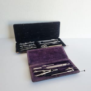 Peut inclure: Deux coffrets de dessin vintage ouverts. Un étui est noir avec un intérieur noir, l'autre est violet avec un intérieur violet. Les ensembles comprennent divers compas, diviseurs et autres outils de dessin de couleur argentée.