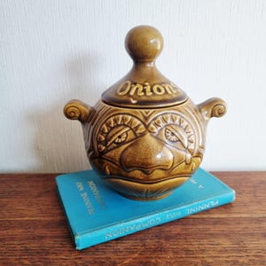 May include: A brown ceramic onion soup tureen with a lid and handles. The tureen has a face design and the word "Onion" on the lid. It sits on a blue book.