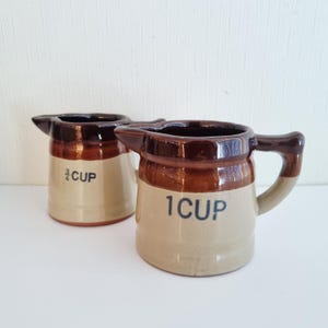 Könnte beinhalten: Zwei Messkrüge aus Keramik. Auf einem Krug steht "3/4 CUP" und auf dem anderen "1 CUP". Die Krüge sind beige mit braunen Akzenten und einem Griff. Die Krüge haben einen Ausguss.