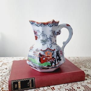 May include: A white ceramic pitcher with a red, blue, and green floral pattern. The pitcher has a octagonal shape and a handle with a floral design. The pitcher is sitting on top of a red book with the title "The Commodore" by C.S. Forester.