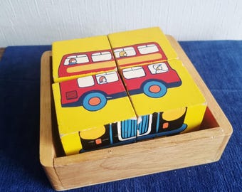 May include: Wooden block puzzle featuring a red double-decker bus design on a yellow background. The puzzle is composed of four blocks and sits in a natural wood tray. The bus has blue wheels and windows with passengers.