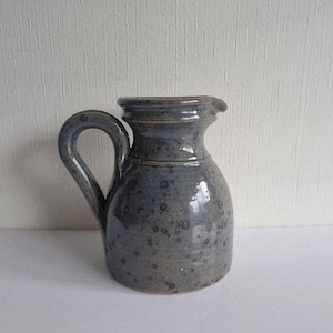 Könnte beinhalten: Handgefertigter Keramikkrug in Blau- und Grautönen. Der Krug hat einen abgerundeten Körper, einen gebogenen Henkel und einen kleinen Ausguss. Die Oberfläche ist strukturiert und mit dunklen Sprenkeln versehen.